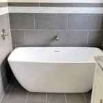 A white bathtub in a gray tiled bathroom with a shower.